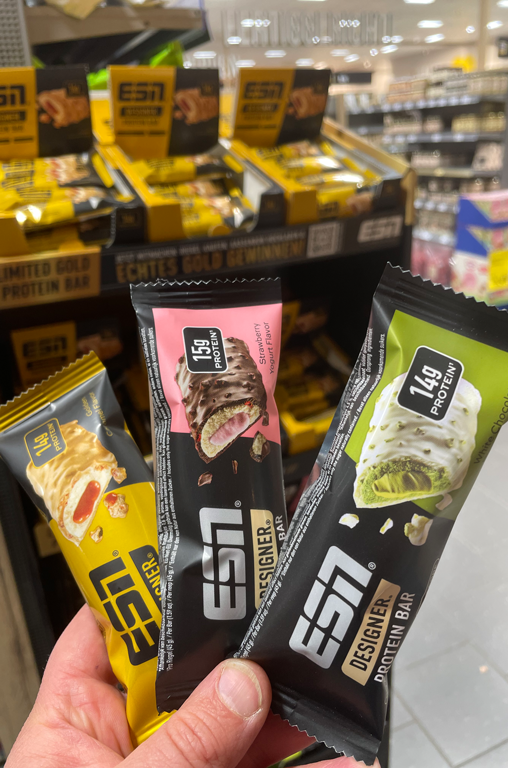 Die drei neuen ESN-Riegel-Sorten "strawberry yoghurt", "golden caramel" und "white chocolate pistachio" im Markt vor dem ESN-Aufsteller fotografiert